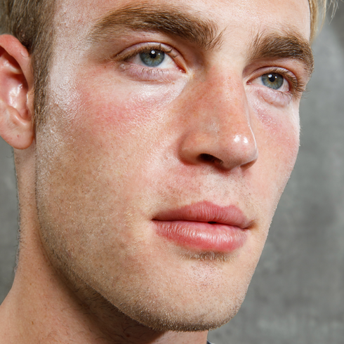 Close-up of a young man's clean-shaven face with blue eyes, natural skin texture, and light facial stubble on gray background.