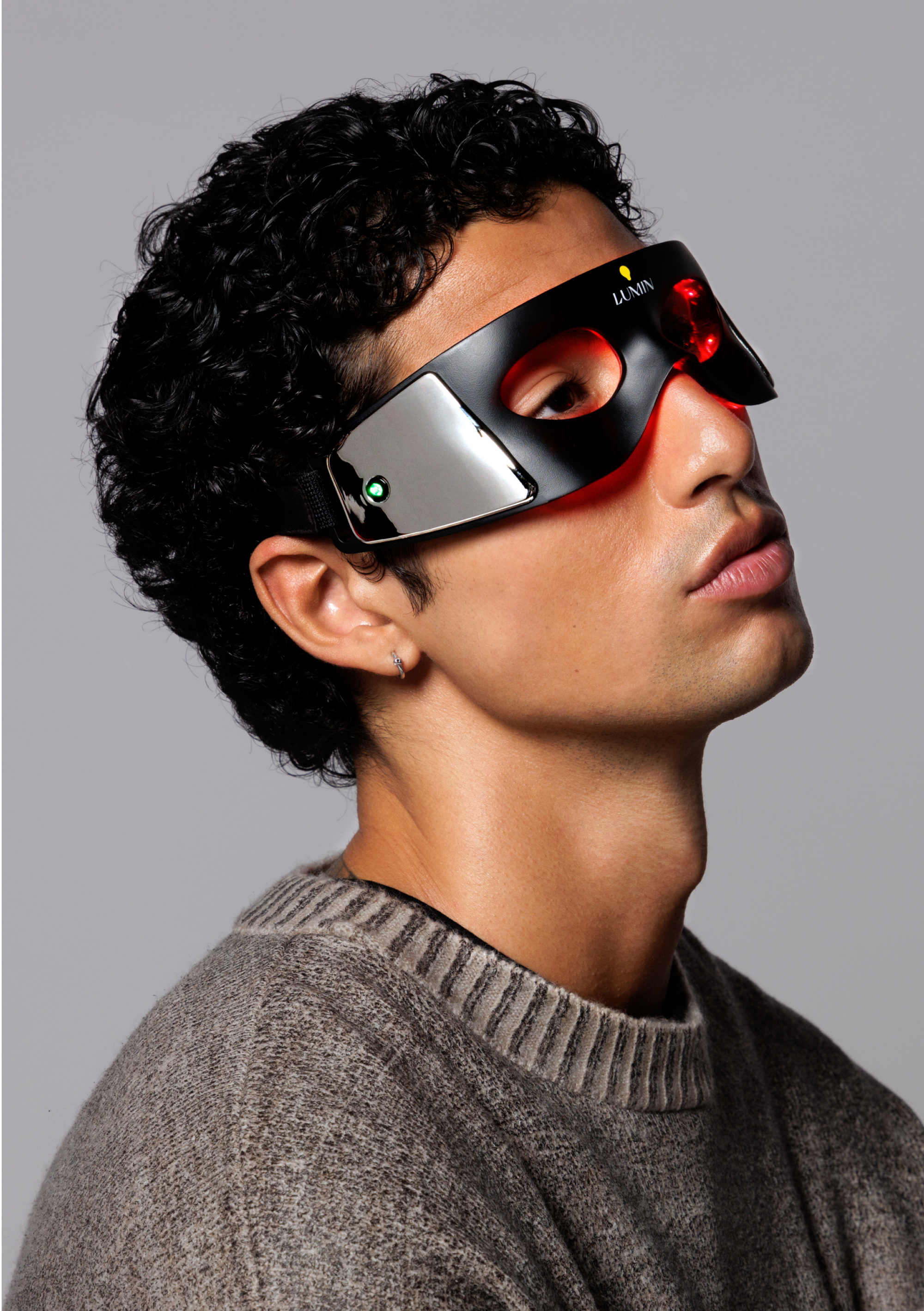 Man wearing black Lumin LED light therapy eye mask with red lights, gray knit sweater, and silver earring against gray background