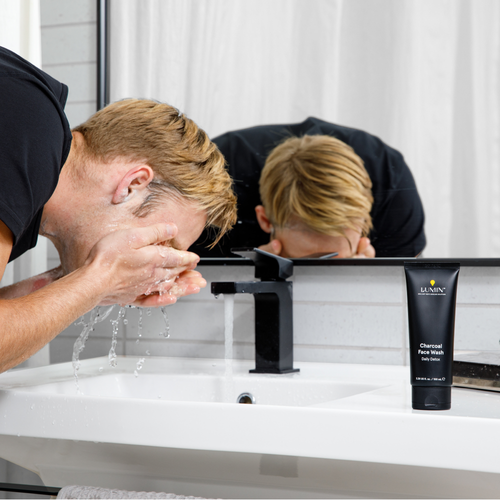 Man washing face with water at bathroom sink next to Lumin charcoal face wash tube for daily detox skincare.