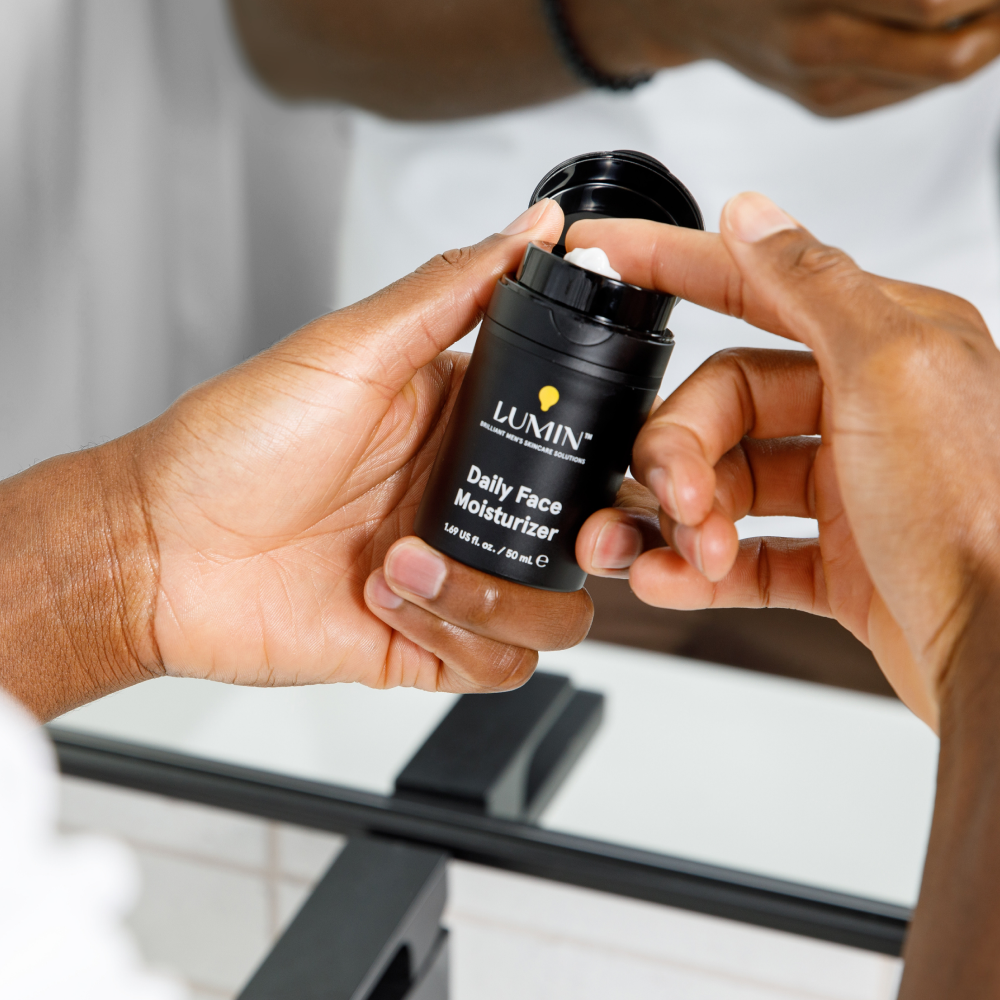 Black Lumin Daily Face Moisturizer bottle held by hands, with cream being scooped, bathroom mirror background.