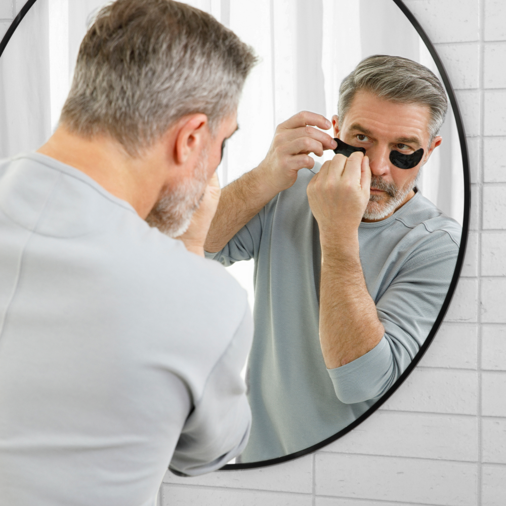 Middle-aged man applying black under eye patches for skincare in bathroom mirror, anti-aging routine.