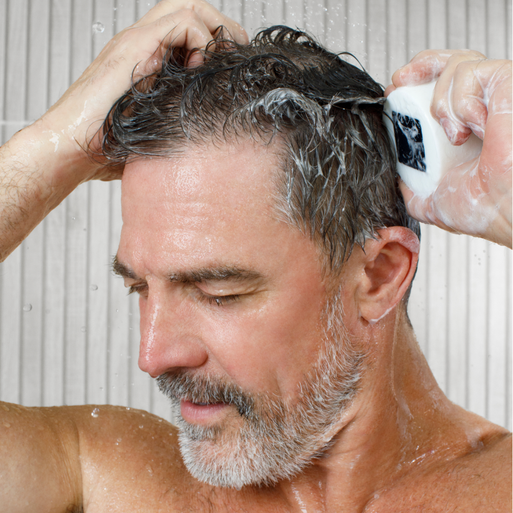 Man washing wet gray hair with white shampoo bar in shower, skincare and haircare routine for men with beard.