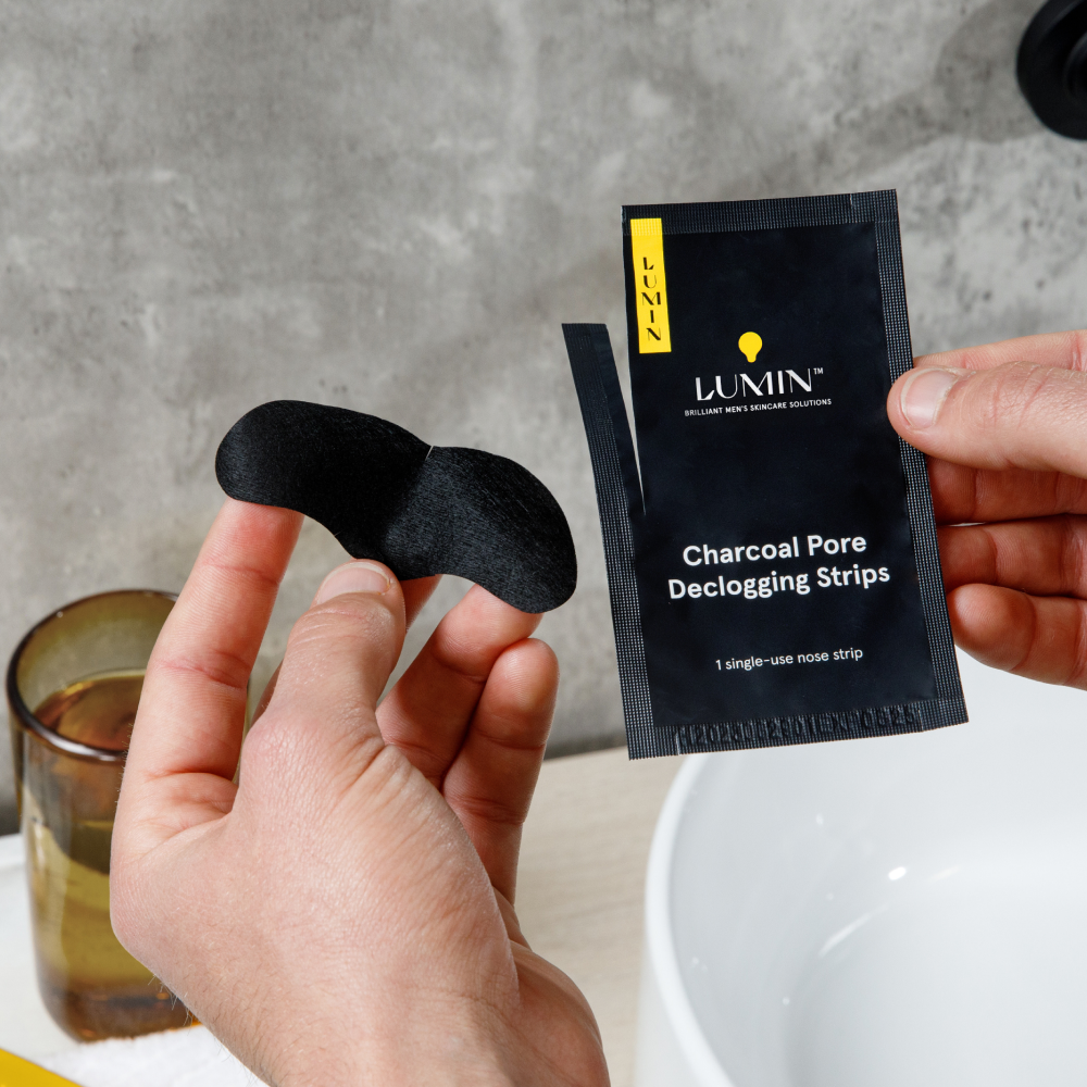 Lumin charcoal pore declogging nose strip held by hands above white sink and brown glass on bathroom counter