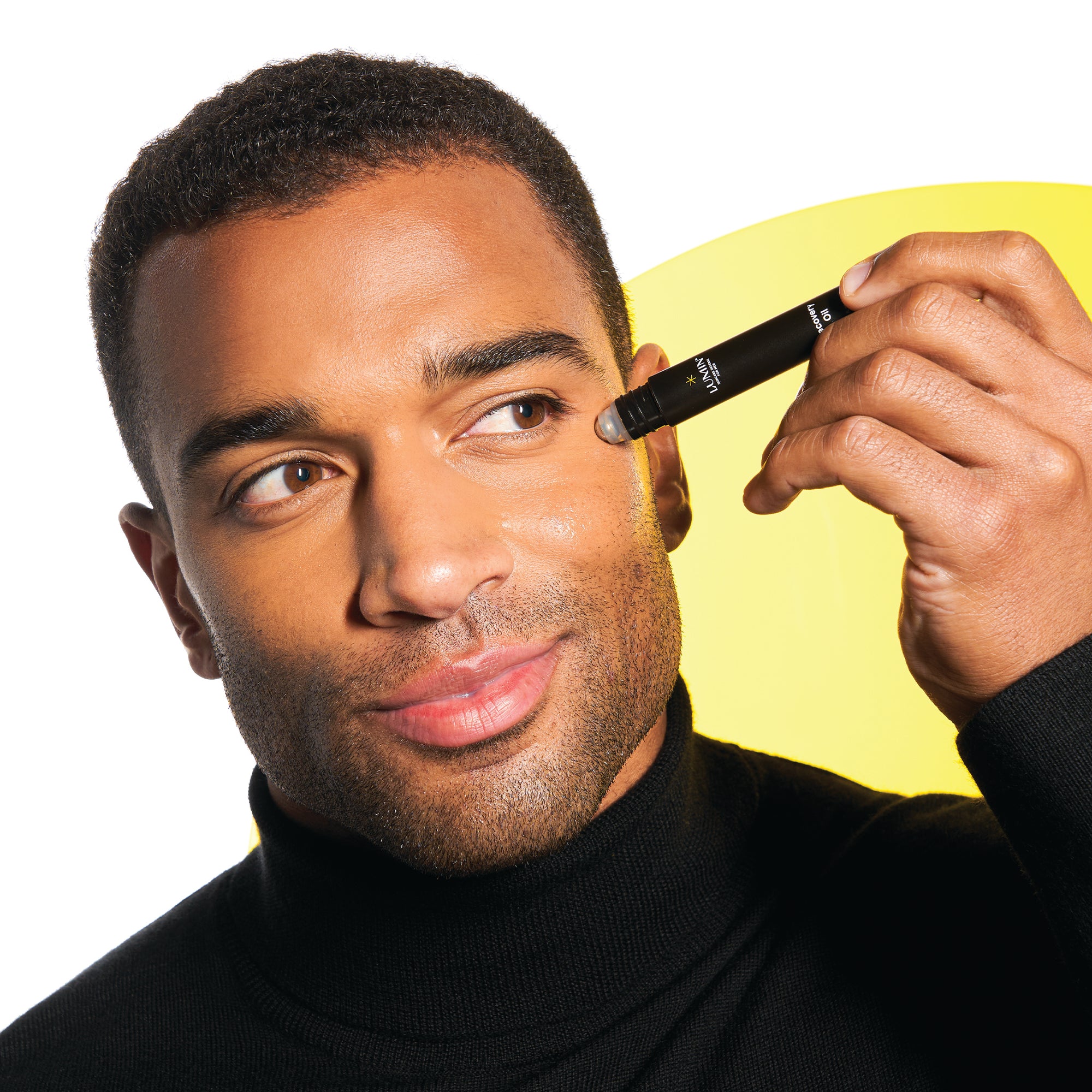 Man applying black rollerball skincare serum near eye, glowing skin, black turtleneck, yellow accent background for beauty care.