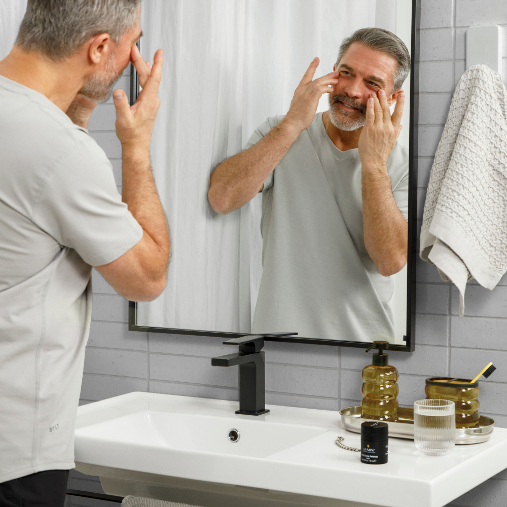 Middle-aged man applying anti-aging cream in modern bathroom mirror with skincare products and black faucet on sink