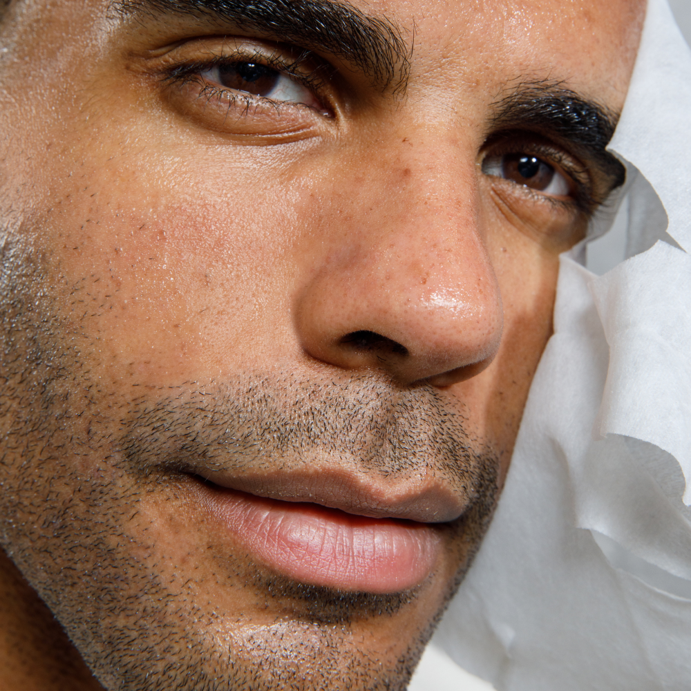 Close-up of a man's face with smooth skin, subtle stubble, and brown eyes, holding a white facial cleansing wipe.